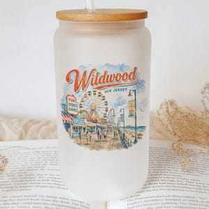 A frosted glass tumbler with a wooden lid, featuring an illustration of a boardwalk and ferris wheel, placed on top of an open book.