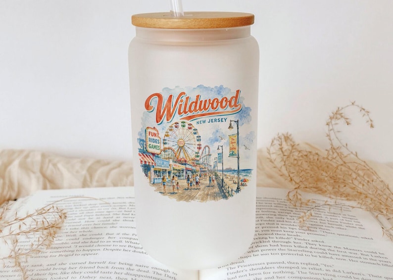 A frosted glass tumbler with a wooden lid, featuring an illustration of a boardwalk and ferris wheel, placed on top of an open book.