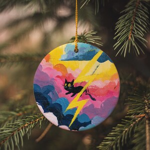 May include: Round Christmas ornament with a painted design of a black cat, yellow lightning bolts, and colorful clouds. The ornament is hanging from a gold string against a backdrop of evergreen branches.