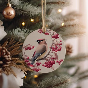 May include: Round ceramic Christmas ornament featuring a bird perched on a branch with red berries. The bird has brown, gray, and red feathers. The ornament hangs from a gold string and is displayed on a Christmas tree.