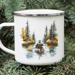 May include: White enamel mug with a watercolor illustration of a person paddling a canoe down a river through a forest. The trees are painted in shades of green, brown, and yellow.