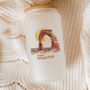 May include: Frosted glass tumbler with a wooden lid, featuring a watercolor illustration of Delicate Arch in Arches National Park. The design includes a yellow sun and the text "Arches National Park".