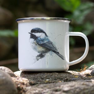 May include: White enamel mug with a silver rim and a large white handle. The mug features a watercolor painting of a small bird with black, white, and blue feathers. The mug is sitting on a rock.
