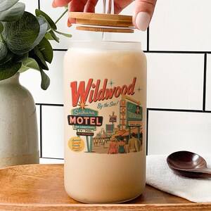 A hand is opening a glass jar with a label that features a vintage-style motel advertisement. The jar is placed on a wooden surface, with a vase of greenery and a wooden spoon nearby.