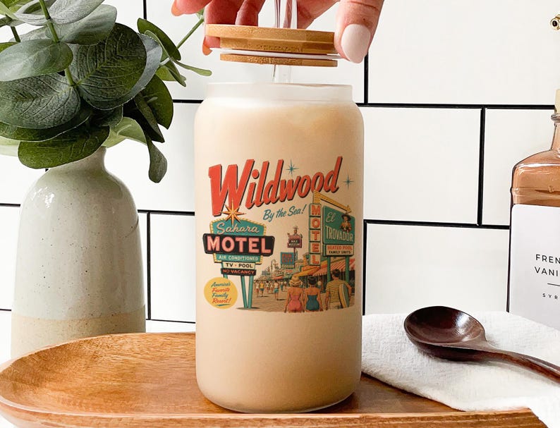 A hand is opening a glass jar with a label that features a vintage-style motel advertisement. The jar is placed on a wooden surface, with a vase of greenery and a wooden spoon nearby.