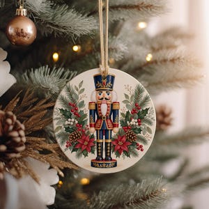May include: A round ceramic Christmas ornament featuring a central illustration of a nutcracker in a blue and red uniform, surrounded by green pine branches, red poinsettia flowers, pinecones, and red berries, suspended by a tan twine loop.