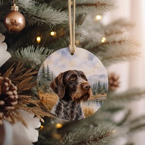 May include: Round ceramic Christmas ornament featuring a detailed portrait of a brown and gray dog with a long snout. The dog is set against a backdrop of a lake, trees, and mountains. The ornament hangs from a gold string.