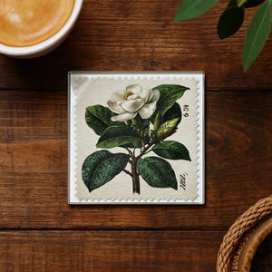 May include: Square coaster featuring a botanical illustration of a white magnolia flower with green leaves and buds. The design is set against a cream-colored background with a postage stamp border. The coaster is on a wooden surface.