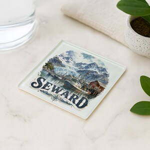 May include: Square glass coaster featuring a scenic illustration of Seward, Alaska, with mountains, water, and a building. The word "SEWARD" is prominently displayed in a vintage font. A compass rose is also visible. The coaster is on a white surface.