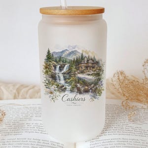 A white glass jar with a wooden lid, featuring a landscape scene with a waterfall and a cabin, sits on top of an open book.