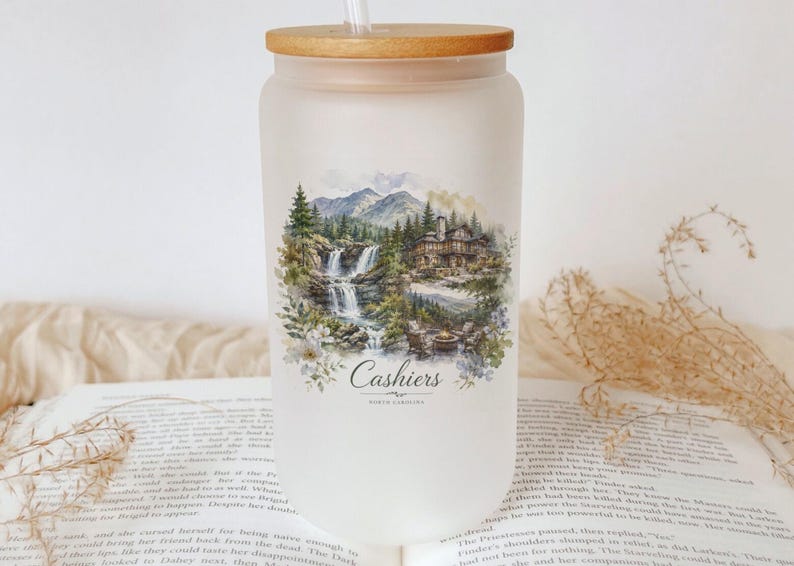 A white glass jar with a wooden lid, featuring a landscape scene with a waterfall and a cabin, sits on top of an open book.