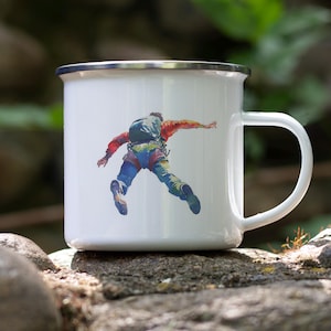 May include: White enamel mug with a silver rim and a white handle. The mug features a colorful illustration of a person in mid-air, wearing climbing gear. The mug is sitting on a rock.