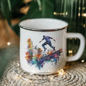May include: White enamel mug with a black rim and a large handle. The mug features a watercolor illustration of a person jumping over a cityscape. The artwork includes buildings and colorful splashes of paint.