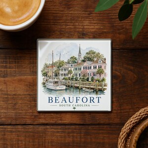 May include: Square coaster featuring a watercolor illustration of Beaufort, South Carolina. The image depicts a waterfront scene with boats, buildings, and the text "BEAUFORT SOUTH CAROLINA". The coaster is set on a dark wood surface.