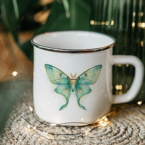 May include: White ceramic mug with a silver rim and a large, detailed illustration of a Luna moth in shades of teal and gold. The mug has a curved handle and is set on a woven mat.