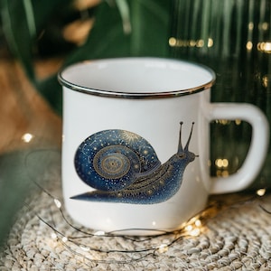 May include: White enamel mug with a black rim and handle. The mug features a detailed illustration of a blue and gold snail with a starry shell. The snail design is centered on the mug's front, creating a whimsical and artistic look.