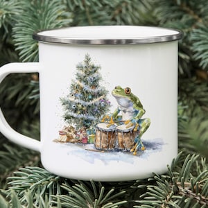 May include: White enamel mug with a silver rim featuring a watercolor illustration of a green frog playing drums next to a decorated Christmas tree and wrapped gifts. The mug has a white handle.