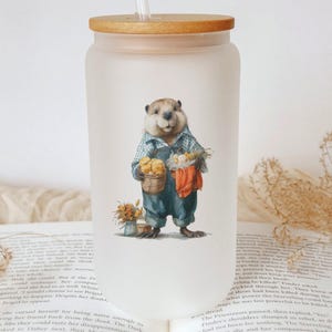 May include: Frosted glass tumbler with a wooden lid and straw. The tumbler features a whimsical illustration of a beaver dressed in overalls, holding baskets of produce. The design includes a small bouquet of flowers.