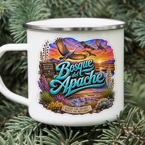 May include: A white enamel mug with a silver rim featuring a colorful graphic of the Bosque del Apache National Wildlife Refuge. The design includes flying cranes, a sunset landscape, desert plants, and the text Bosque del Apache.
