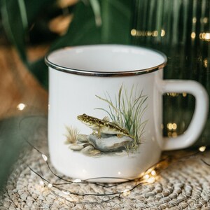 May include: White enamel mug with a silver rim, featuring a detailed illustration of a fish on a rock with tall grass. The mug has a large handle and is set against a blurred background with decorative lights.