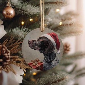 May include: A round Christmas ornament featuring a German Shorthaired Pointer wearing a Santa hat. The dog is brown and white with a red gift box. The ornament hangs from a gold ribbon and is displayed on a Christmas tree.