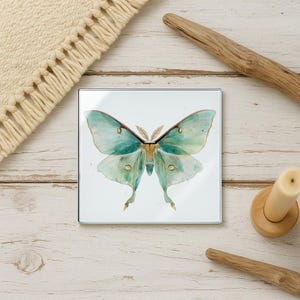 May include: Square glass coaster featuring a watercolor illustration of a Luna moth. The moth has teal and green wings with delicate details. The coaster is set against a white background.