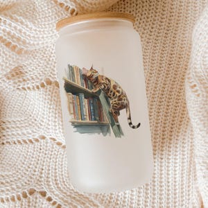 May include: Frosted glass tumbler with a bamboo lid. The tumbler features a watercolor illustration of a Bengal cat climbing a bookshelf filled with colorful books. The design is set against a soft, neutral background.