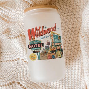 A white candle jar with a vintage-style label that reads "Wildwood by the Sea" and features an illustration of a motel and people.