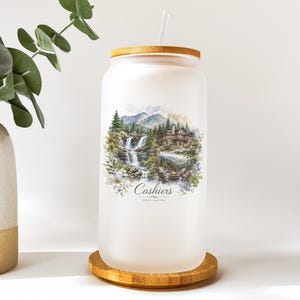 A frosted glass jar with a wooden lid, featuring a landscape scene with a waterfall and mountains, and the text "Cashiers NORTH CAROLINA" printed on it.