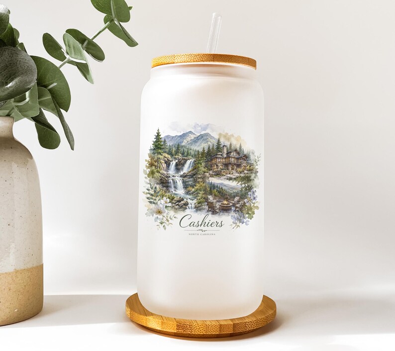 A frosted glass jar with a wooden lid, featuring a landscape scene with a waterfall and mountains, and the text "Cashiers NORTH CAROLINA" printed on it.