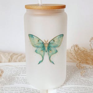 May include: Frosted glass tumbler with a wooden lid and a straw. The tumbler features a watercolor illustration of a blue and green moth. The design is centered on the front of the glass. The tumbler is sitting on top of an open book.