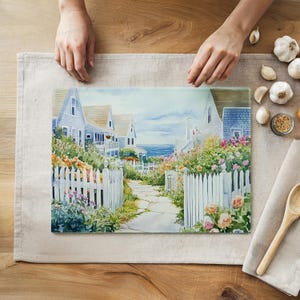 May include: A glass cutting board featuring a watercolor painting of a coastal scene. The artwork depicts light blue houses with white picket fences, a stone path, and colorful flowers. The ocean is visible in the background.