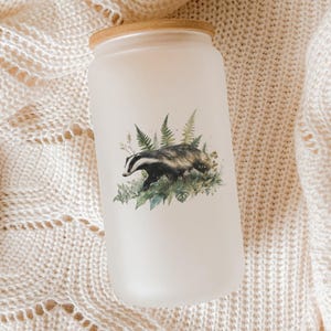 May include: A frosted glass tumbler with a bamboo lid, featuring a watercolor illustration of a badger surrounded by ferns and foliage. The tumbler is cylindrical and rests on a cream-colored knitted surface.