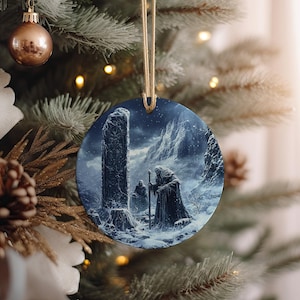 May include: Round Christmas ornament featuring a winter landscape with a robed figure holding a staff, standing before a tall stone structure. The ornament is hanging from a gold string, with a gold bauble and pine needles in the background.
