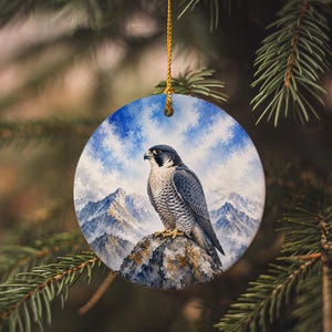 May include: Round ceramic ornament featuring a detailed painting of a falcon perched on a rock, set against a backdrop of snow-capped mountains and a blue sky with clouds. The ornament is suspended by a gold string.