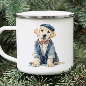 May include: White enamel mug featuring a watercolor illustration of a Labrador puppy dressed in a blue blazer, bow tie, and cap. The mug has a silver rim and a white handle. The background is blurred evergreen branches.
