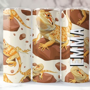 May include: A white tumbler with a pattern of orange and yellow bearded dragons on a beige background. The text "EMMA" is printed on the tumbler.