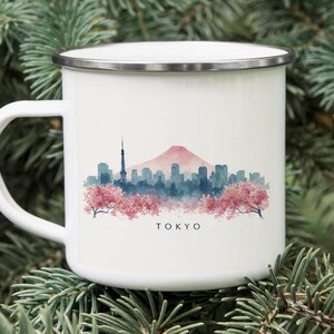 May include: White enamel mug with a silver rim, featuring a watercolor illustration of the Tokyo skyline. The design includes a pink mountain, blue buildings, and cherry blossoms. The word "TOKYO" is printed below the image.