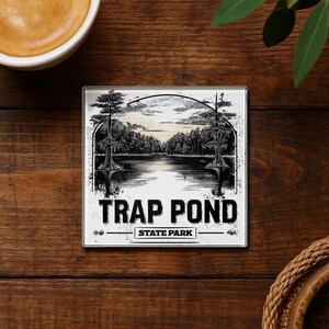May include: Square coaster with a black and white illustration of a lake scene with trees and the text "TRAP POND STATE PARK". The coaster has a white border and is set on a dark wood surface.