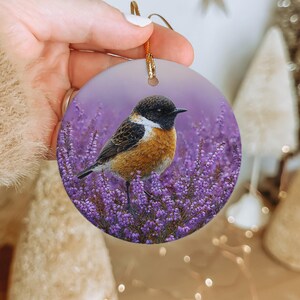 May include: Round ceramic ornament featuring a detailed illustration of a small bird with orange, black, and white plumage perched amidst purple flowers. The ornament has a gold string for hanging and a soft purple background.
