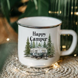 May include: White enamel mug with a black rim and handle. The mug features the text "Happy Camper" above an illustration of a vintage camper trailer surrounded by pine trees. The mug is on a woven surface.