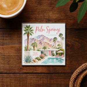 May include: Square coaster featuring a watercolor illustration of Palm Springs, California. The design includes palm trees, mountains, a swimming pool, and the text "Palm Springs" in pink. The coaster has a silver border and is set on a wooden surface.