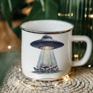 May include: White enamel mug with a black rim and handle. The mug features a whimsical illustration of a UFO beaming up a herd of cows. The UFO is dark blue, and the cows are black and white.