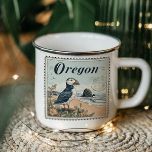 May include: White ceramic mug with a black rim and a handle. The mug features a vintage-style illustration of a puffin bird on a beach with the word "Oregon" above. The mug is on a woven surface.