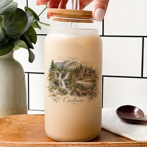 a hand opening a jar with a landscape scene printed on it, placed on a wooden surface. There is also a wooden spoon and a bottle of vanilla-scented liquid nearby.