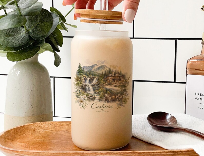 a hand opening a jar with a landscape scene printed on it, placed on a wooden surface. There is also a wooden spoon and a bottle of vanilla-scented liquid nearby.
