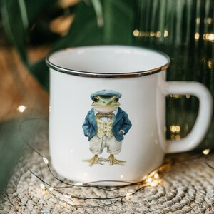 May include: White enamel mug with a black rim featuring a whimsical illustration of a frog in a blue blazer, vest, shorts, and cap. The frog has a bow tie and is standing on its hind legs. The mug has a large handle.