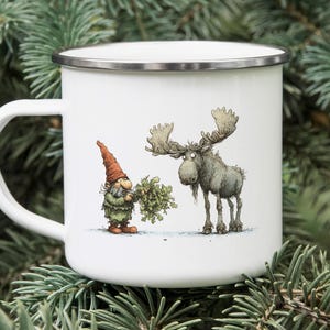 May include: White enamel mug featuring a whimsical illustration of a gnome in a green outfit and a moose. The gnome holds a bundle of greenery, and the moose has large antlers. The mug has a silver rim and a white handle.