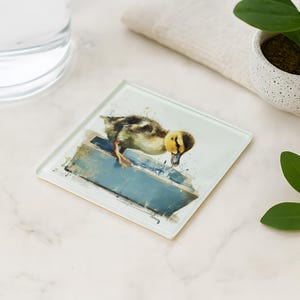 May include: Square glass coaster featuring a watercolor painting of a yellow and brown duckling. The duckling is perched on a blue structure. A glass of water, a small potted plant, and a folded cloth are in the background.