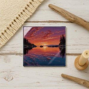 May include: Square glass coaster featuring a scenic sunset over a lake. The image shows a boat with two figures, silhouetted against the vibrant orange and purple sky. The coaster is surrounded by a wooden candle holder, a piece of driftwood, and a cream-colored woven mat.